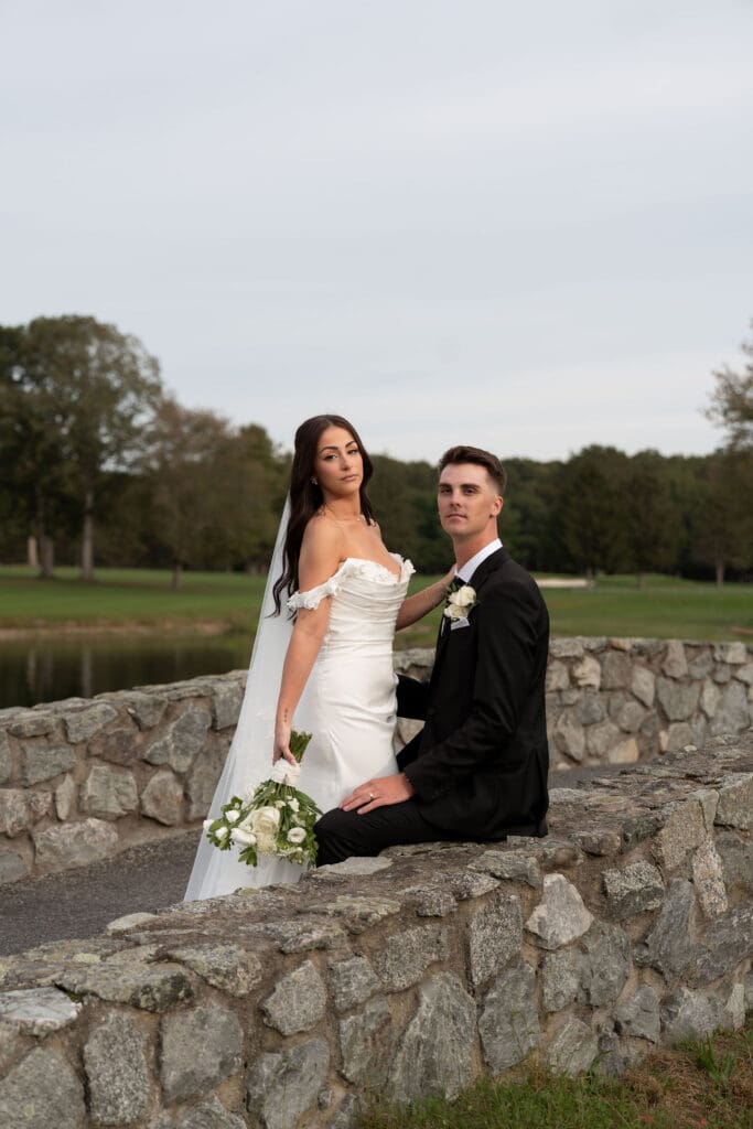 The bride stands beside the seated groom on a low stone wall overlooking the grounds in a calm, classic portrait by CT wedding photographers.