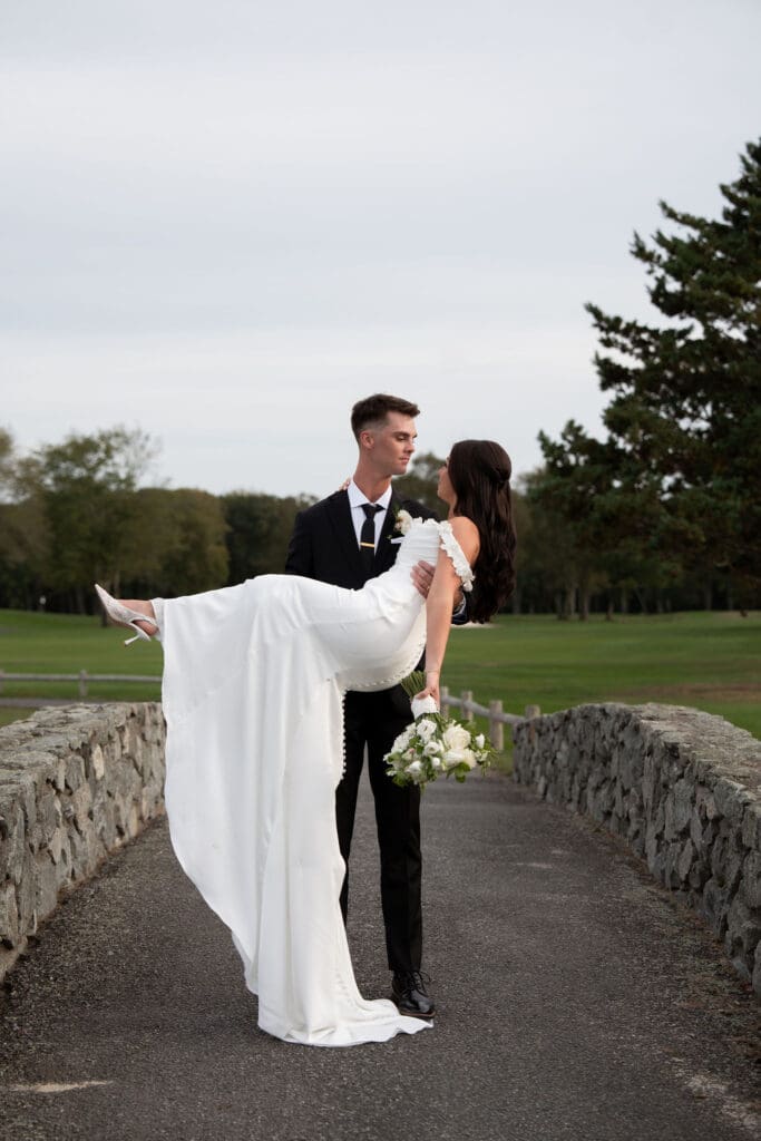 On a stone path beside the lawn, the groom lifts the bride in his arms for a playful portrait from a wedding photographer CT gallery.