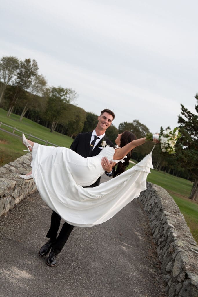 Smiling on the path, the groom lifts the bride in his arms with the grounds stretching behind them, a playful moment from wedding photographers in CT.