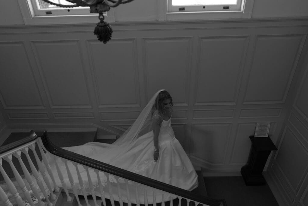 A quiet black-and-white portrait shows the bride descending the staircase with her veil trailing behind her, captured during an Eolia Mansion wedding.