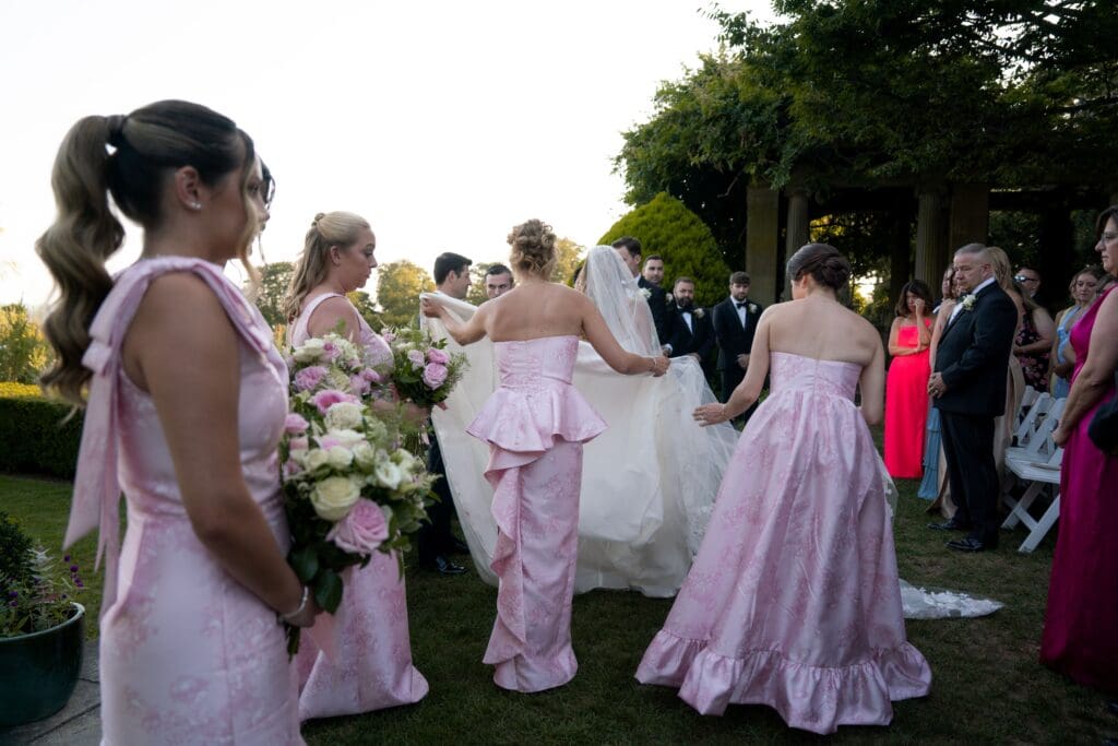 Bridesmaids in blush dresses gather around the bride on the lawn in a joyful pre-ceremony moment from a Connecticut wedding venues outdoor guide.