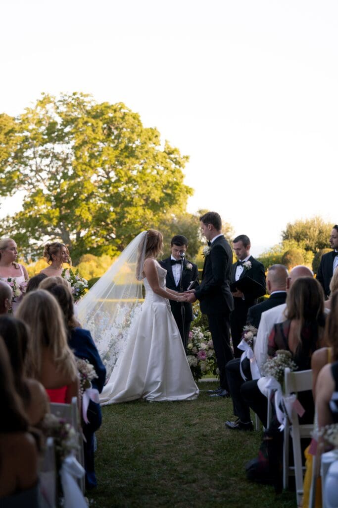 The couple stands at the altar during an outdoor ceremony as guests line both sides of the lawn, photographed at one of the most elegant Connecticut wedding venues outdoor.
