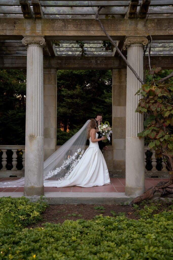 Beneath the estate pergola, the bride stands with her bouquet and veil flowing behind her during a romantic Eolia Mansion wedding.