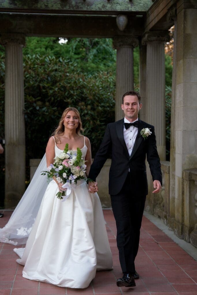 The bride and groom walk hand in hand beneath the pergola, framed by stone columns and greenery at one of the most timeless outdoor wedding venues in Connecticut.