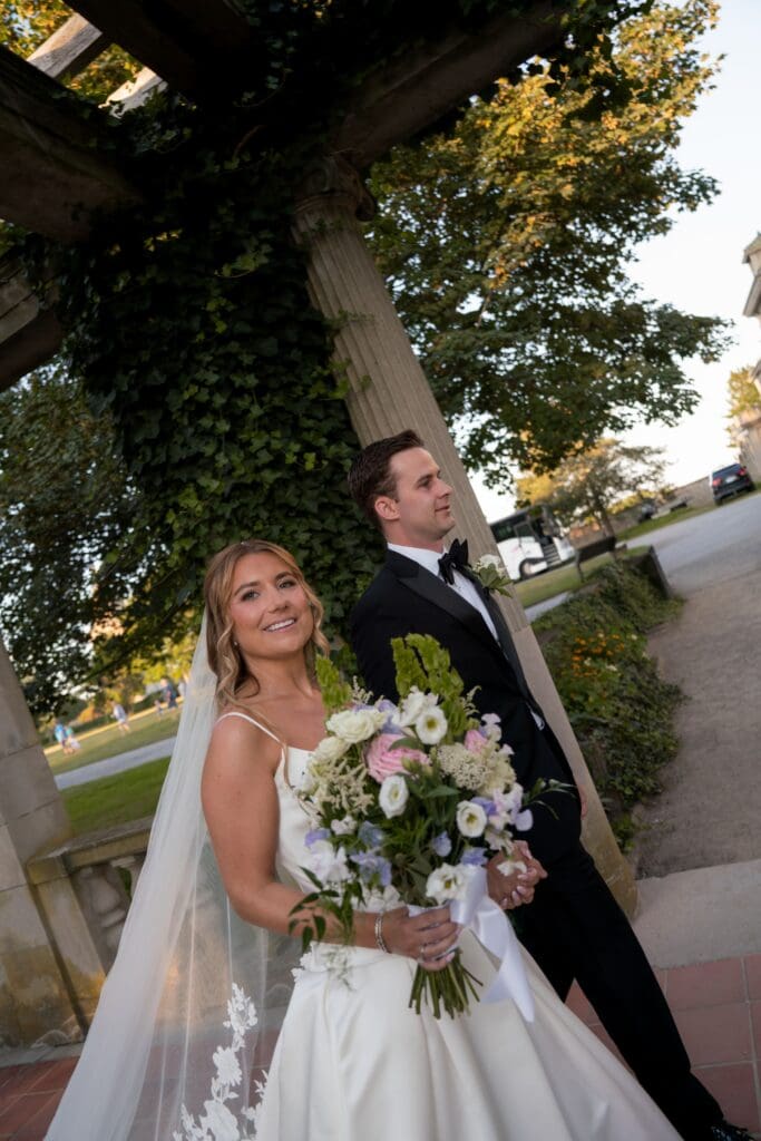 The bride smiles toward the camera with bouquet in hand while the groom stands beside her under the vine-covered pergola, from a mansion wedding photographer Connecticut gallery.