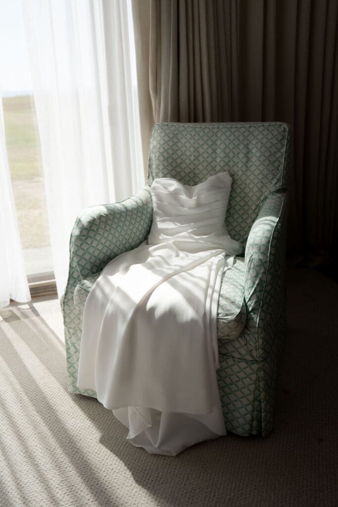 Sunlight falls across the bride’s dress draped over an upholstered chair, creating a quiet detail image for an estate wedding photographer in Connecticut.