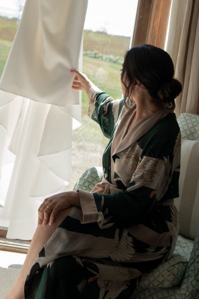 A bridesmaid sits by the window holding the wedding dress as it catches the light, captured in an editorial moment by a Connecticut estate wedding photographer.