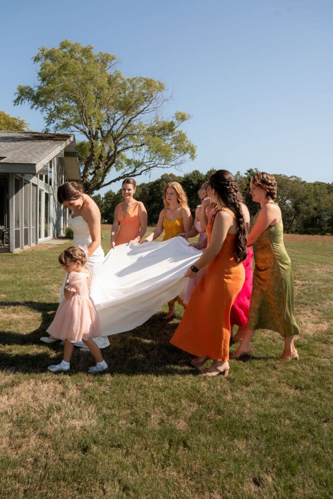Bridesmaids gather around the bride outdoors, lifting the skirt of her gown in a playful moment photographed by a CT wedding photographer.
