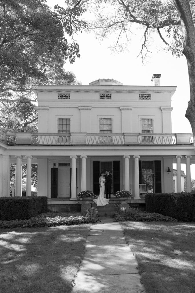A black-and-white wide shot frames the mansion façade with the couple standing beneath the columns, capturing the scale and elegance of the estate with a Wedding Photographer CT perspective.
