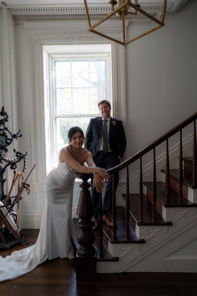 The bride leans on the staircase banister while the groom stands above her in soft window light, a refined interior portrait by a Connecticut wedding photographer.