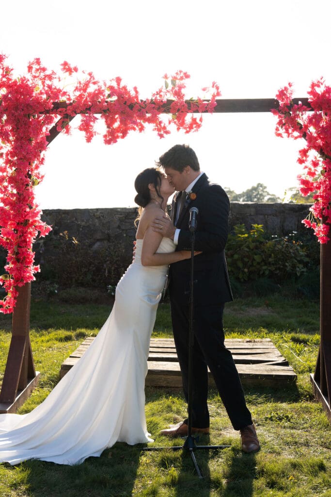The couple kisses beneath a floral ceremony arch as sunlight filters through the garden, a moment from a Connecticut wedding venues guide focused on timeless celebrations.