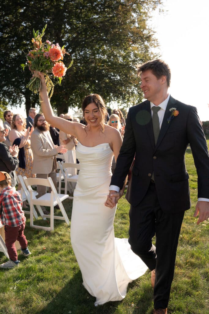 Just married, the couple walks back up the aisle smiling as guests cheer in golden light at one of the most beautiful outdoor wedding venues in Connecticut.