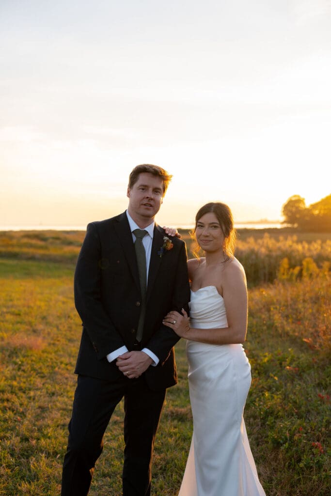 The couple stands together in golden light as the sun drops behind the grasses, photographed at a timeless Connecticut wedding venue.