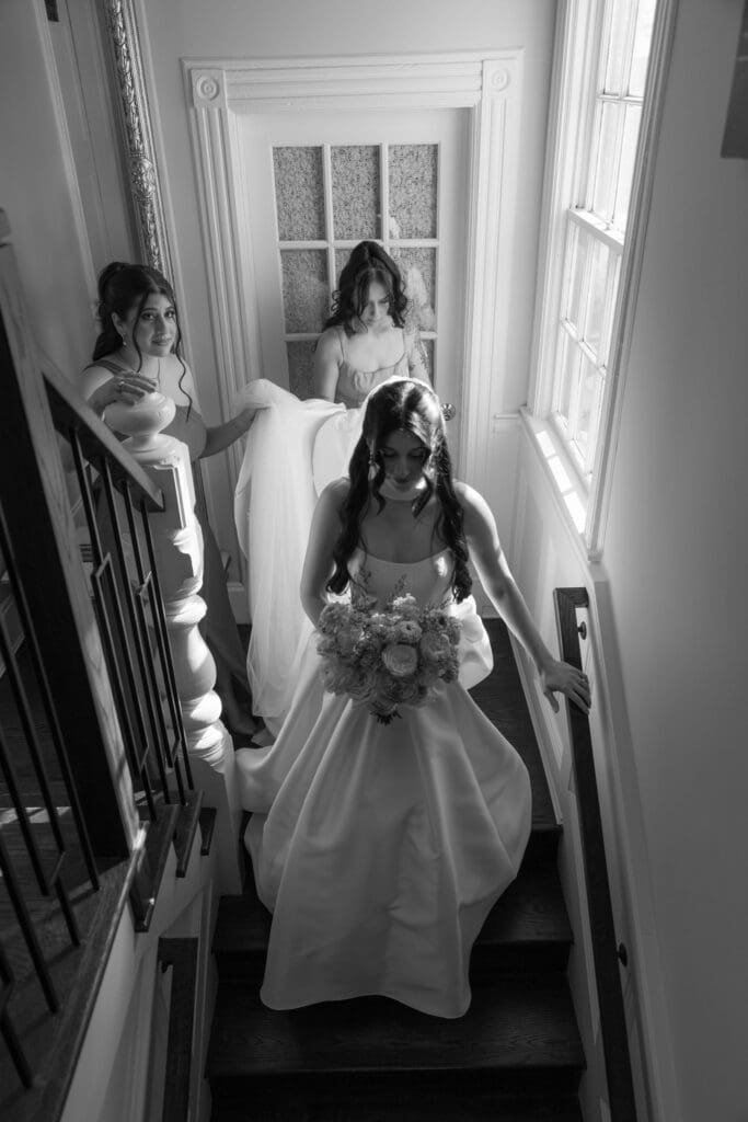 In a black-and-white stairwell portrait, the bride descends with her bouquet while two women follow behind in soft window light, photographed by CT wedding photographers.