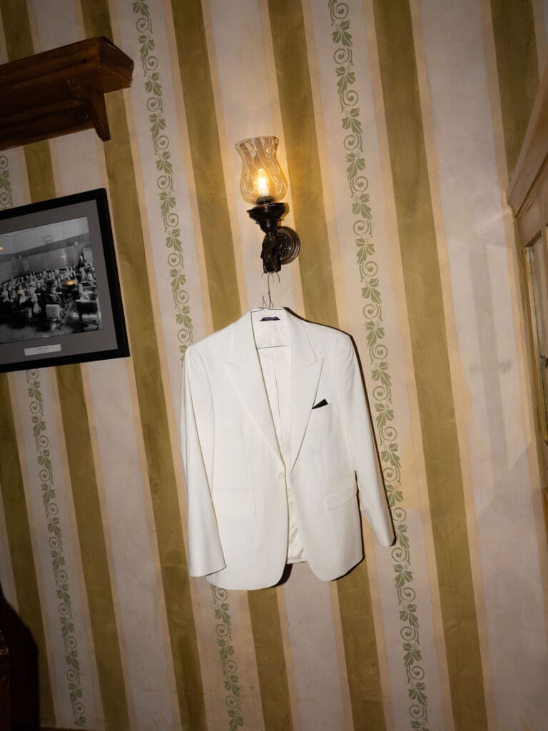 A white dinner jacket hangs beneath a wall sconce and striped wallpaper in a quiet getting-ready detail from a wedding photographer in Connecticut feature.