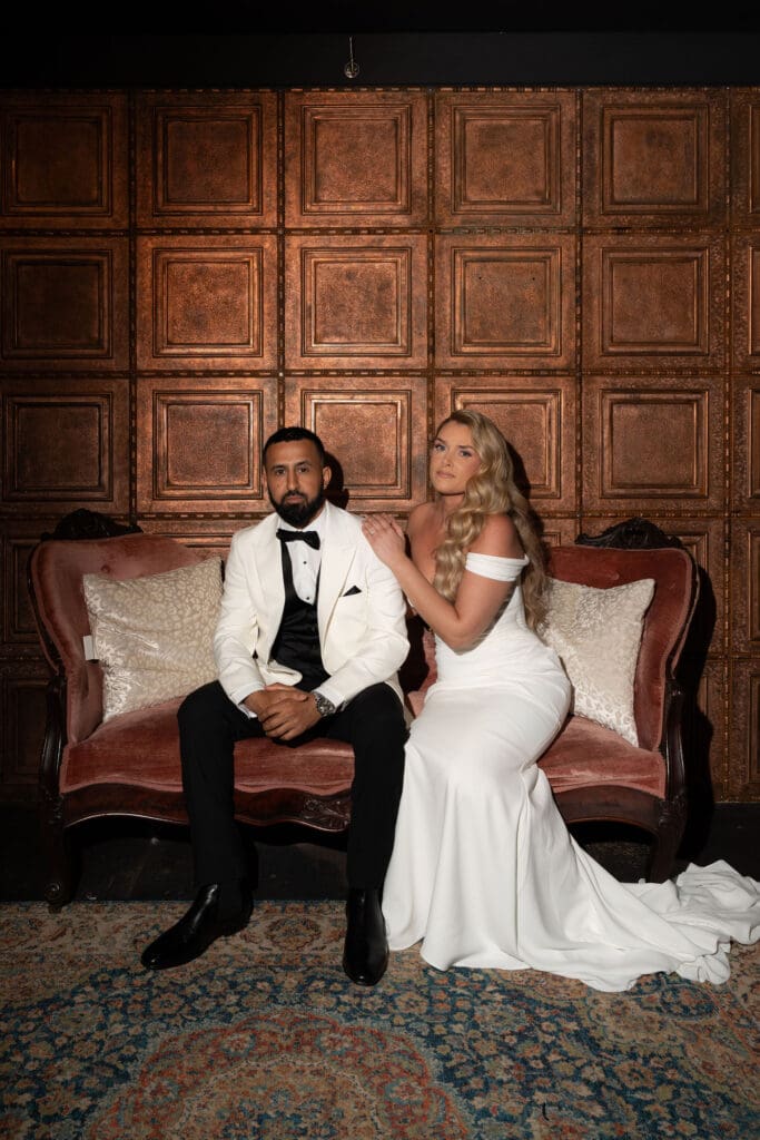 The couple sits together on a velvet settee against carved wood paneling, creating a dramatic portrait with an editorial feel by a wedding photographer in Connecticut.
