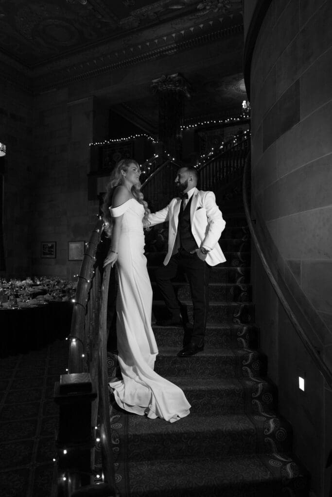In a black-and-white stairwell moment, the bride reaches for the groom as string lights trace the railing in a scene photographed by a Connecticut wedding photographer.