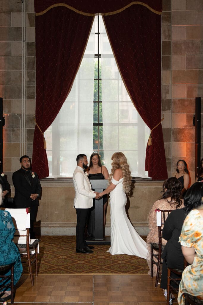 Standing before towering windows and deep red drapery, the couple exchanges vows in a ceremony space that feels timeless and refined at a historic wedding venue in Connecticut.
