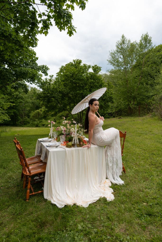Seated at the end of the table with a parasol, the bride is framed by flowers and woodland surroundings in a scene suited to a fine art wedding photographer in Connecticut.