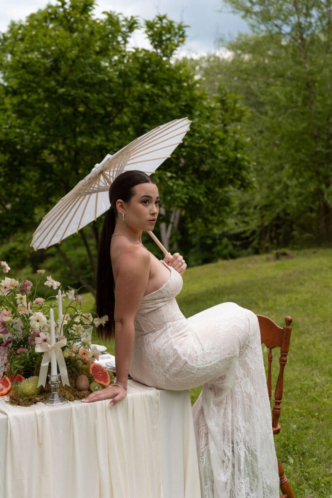 Seated with a parasol beside the floral table, the bride is photographed in gentle natural light by a true to color wedding photographer Connecticut.