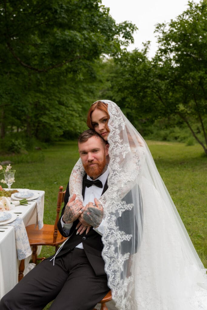 The bride drapes her veil over the seated groom beside an outdoor tablescape, creating an intimate editorial portrait by a romantic wedding photographer Connecticut couples love.
