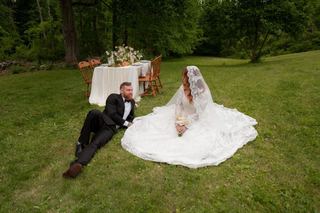 Bride and groom sit together on the grass beside a candlelit reception table set in the woods, creating an intimate editorial portrait by a Connecticut wedding photographer.