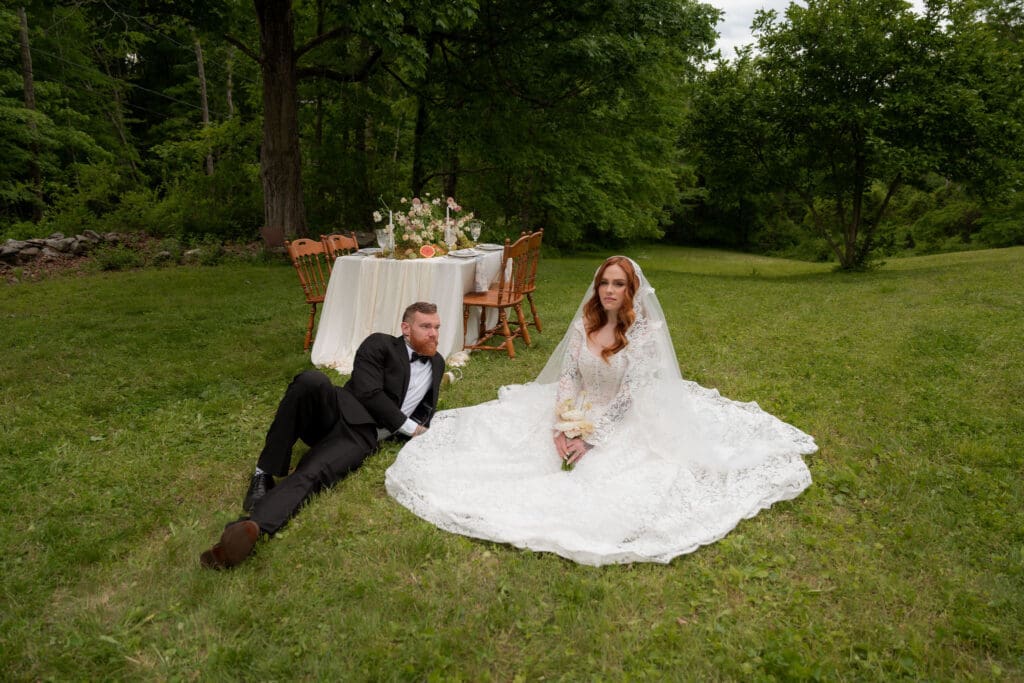 The bride sits on the grass while the groom reclines nearby beside an outdoor reception table, creating a relaxed portrait from wedding photographers in CT.