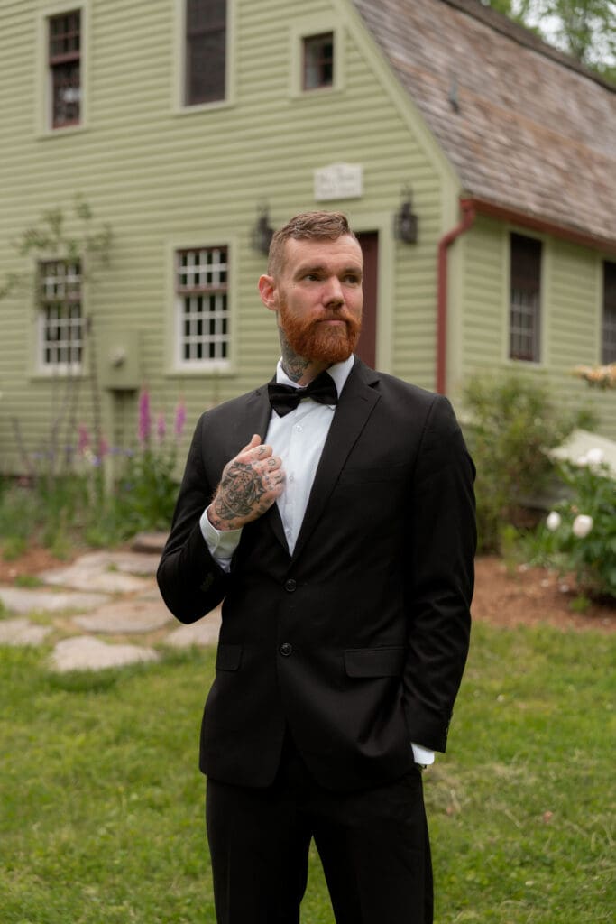 The groom stands in front of a historic house adjusting his bow tie in a classic portrait from a Connecticut wedding photographer session.